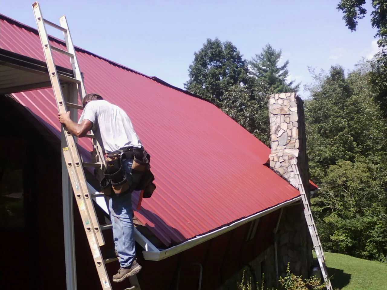 Roofer on a ladder beside a red metal roof and stone chimney — Soffit & Fascia Repair by Roofing Brothers in Murrieta